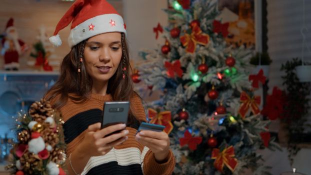 Happy woman looking for christmas gifts at decorated home. Young adult wearing santa hat using smartphone and credit card to shop online for seasonal festive celebration dinner party