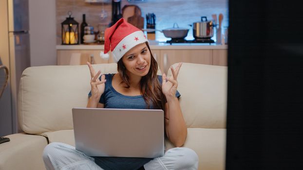 Person talking to family on video call conference to celebrate christmas eve. Woman wearing santa hat while using online conference for remote holiday festivity on laptop in festive room