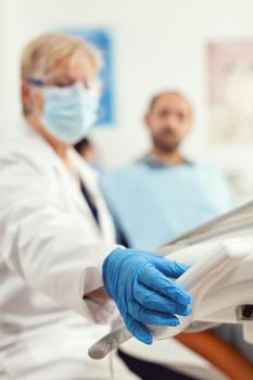 Closeup of stomatologist doctor preparing dental tools for stomatology surgery during dentistry appointment. Orthodontist medic wearing face mask discussing with man patient for medical treatment