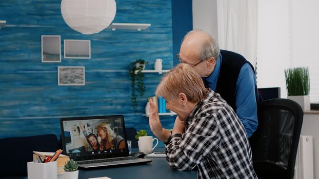 Happy senior couple waving during video conference with nephews using laptop in living room. Elderly person using internet online chat technology video webcam making virtual meeting video call