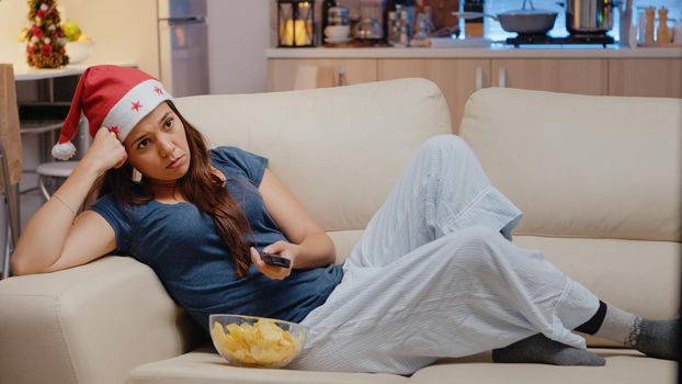 Tired woman switching channels and watching television on christmas eve festivity. Person with red santa hat holding TV remote control, laying on sofa and eating chips from bowl.