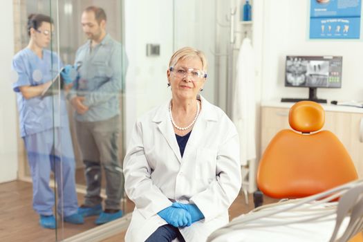 Senior stomatological woman looking into camera while sitting on dental chair in hospital stomatology room. Medical nurse discussing with patient during writing healdcare treatment