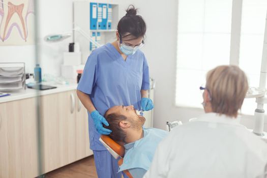 Medical assistant woman doing dental treatment to sick man preparing stomatology tools for dental surgery. Man siting on dentistry chair in hospital orthodontic office during medical procedure