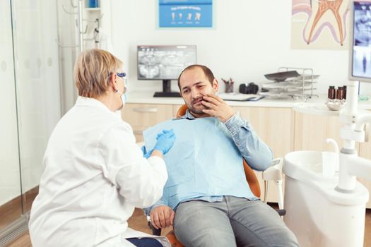 Sick man complaining about tooth pain discussing with stomatologist senior woman about stomatology treatment. Orthodontic doctor preparing for dentistry surgery during medical appointment