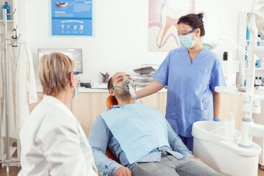 Hospital assistant putting oxigen mask to sick man patient after stomatology surgery, sitting on dental chair in orthodontic hospital room during medical consultation Dentist doctor examining toohache