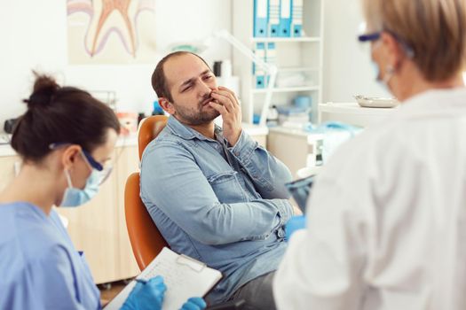 Sick patient with toothache speaking with stomatologist showing affected mass. Man explaining dental problem to doctor while nurse writing stomatological treatment in modern private clinic