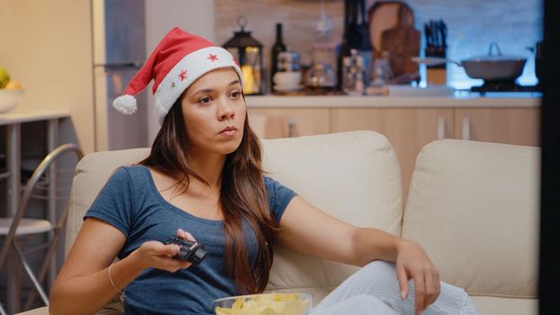 Woman with santa hat watching television on christmas eve. Festive person switching channels on TV with remote control, eating chips from bowl on couch. Adult in holiday season