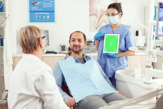 Nurse dentist showing green screen display to stomatology senior doctor while examining tooth pain to man patient sittinh on dental chair. Woman examinating using mock up chroma key tablet