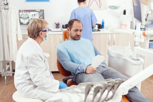 Man patient looking in mirror after medical stomatological team finising dental surgery during stomatology procedure. Senior doctor explaining healthcare treatment for toothache pain