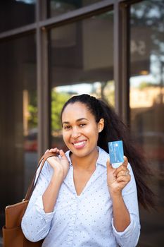 A beautiful mixed race African American woman in a blue collared shirt holds up a blue plastic credit card with micro chip with her brown purse slung over a shoulder outside in front of a storefront.