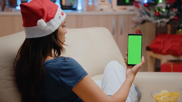 Woman vertically holding smartphone with green screen on christmas eve. Person looking at mockup template and isolated background on cell phone device with chroma key. Adult with gadget