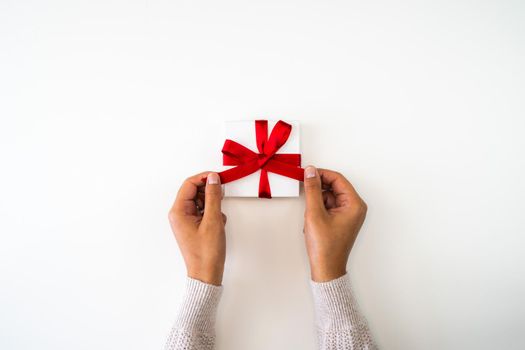 Holiday or Christmas background or backdrop image close up of hands from a mixed race African American woman wearing a sweater tying a red ribbon bow onto a small white gift box with copy space.