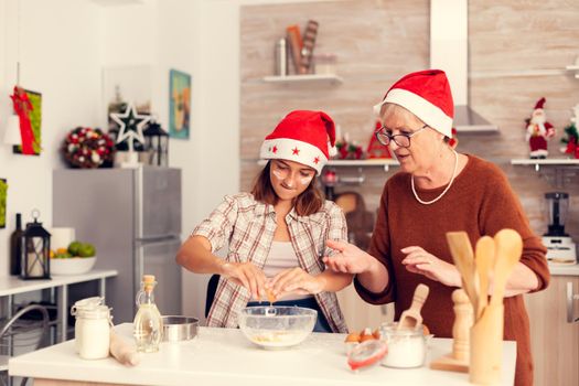 Little niece during christmas day cooking and having fun with grandmother. Happy cheerful joyfull teenage girl helping senior woman preparing sweet cookies to celebrate holidays wearing santa hat.