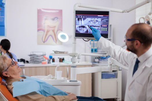 Dentist discussing with senior woman in cabinet holding tablet pc with copy sapce available. Medical teeth care using digital device with patient radiography on tablet pc near patient standing up.