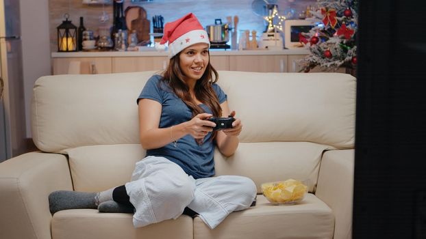 Woman playing video games on console with controller for christmas eve festivity. Adult with santa hat smiling and holding joystick for play on television. Cheerful person on holiday