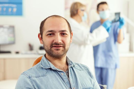 Closeup of patient man looking into camera waiting for doctors to start stomatology surgery sitting on dental chair. Senior doctor and medical nurse examining teeth radiography