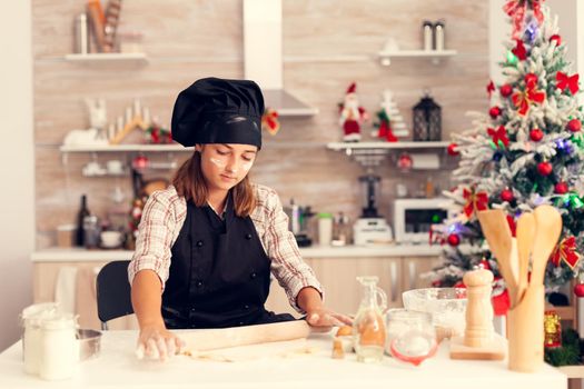 Kid on christmas day making tasty dessert using dough wearing apron. Cheerful happy cute girl while prepearing delicious cookies for christmas celebration in kitchen with christmas tree in the background.