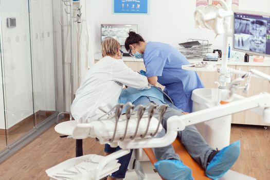 Sick man sitting in dental chair with open mouth for exemination, in stomatological office, doctor preparing for oral problems. Dentist senior woman and medical nurse with masks removes tooth