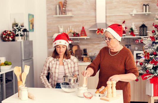 Child learning to make traditional cookies on christmas from grandmother. Happy cheerful joyfull teenage girl helping senior woman preparing sweet cookies to celebrate winter holidays wearing santa hat.