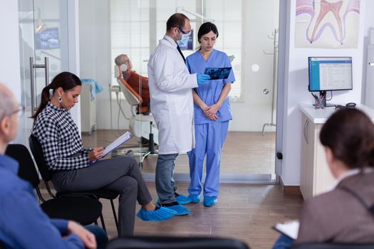 Dentist finishing teeth treatment for old man analysing x-ray radiography with nurse in stomatology waiting room full with patients. People filling form in dentistiry hallway.