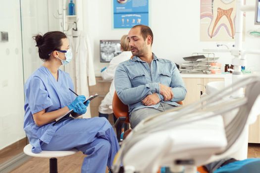 Orthodontist nurse discussing with man patient about tooth consultation writing healthcare treatment on clipboard sitting in medical dental office. Stomatologist making surgery appointment