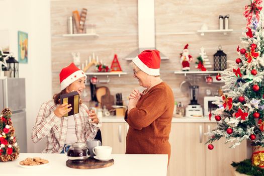 Granny smiling at niece after giving christmas gift box in beautiful decorated kitchen. Senior woman wearing santa hat surprising granddaughter with winter holiday present in home kitchen with xmas tree in the background.