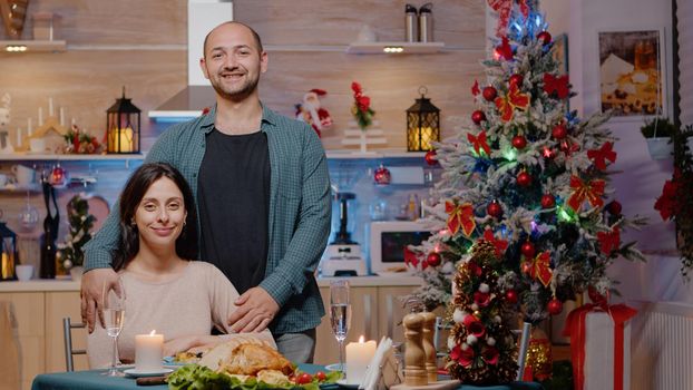 Portrait of couple at festive dinner on christmas eve. Man and woman looking at camera while enjoying holiday meal and celebration, sitting together and feeling cheerful for winter festivity