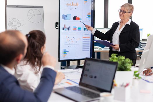 Elderly project manager pointing at desktop presenting statistical data, briefing diverse group of employees. Multiethnical businesspeople working in professional startup financial office during conference meeting
