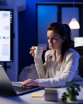 Freelancer woman with a cup of coffee tapping on business computer overworking at marketing project. Busy entrepreneur using modern technology network wireless late at night in company office
