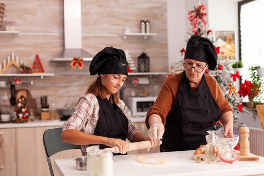 Happy family with apron preparing homemade dough cooking xmas gingerbread dessert in x-mas decorated kitchen. Granddaughter using baking rolling pin making delicious cookies enjoying christmas season