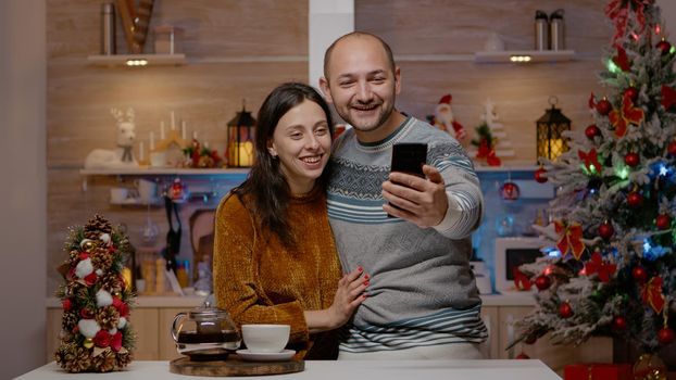 Cheerful couple using video call communication on smartphone talking to family on christmas eve day. Man and woman with remote conversation on online conference for holiday season