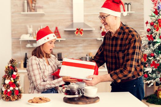 Smiling niece celebrating christmas receiving present box with red bow from grandfather. Senior man wearing santa hat surprising granddaughter with winter holiday gift in home kitchen with xmas tree in the background.