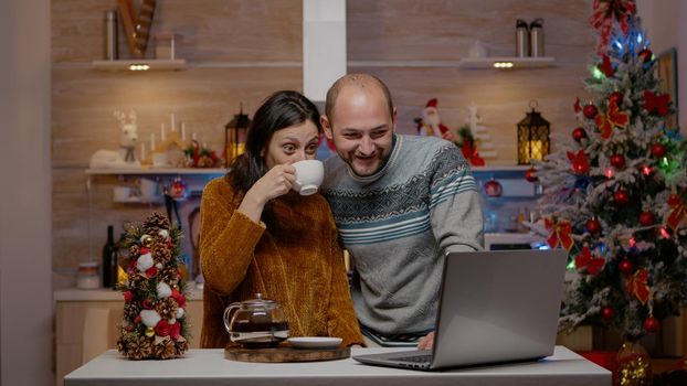 Festive couple using video call communication on laptop celebrating christmas eve in decorated kitchen. Cheerful people talking to family on online conference for holiday season festivity