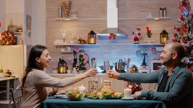 Couple raising glasses of champagne celebrating christmas eve. Festive man and woman sitting at table eating chicken and drinking alcohol for traditional dinner on seasonal holiday.