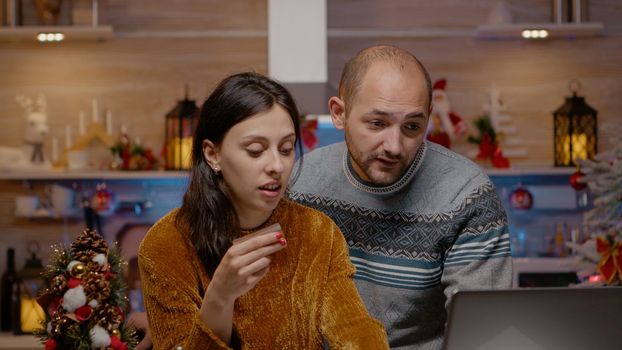 Festive couple doing christmas shopping online with credit card, buying presents for relatives and friends. Man and woman using card and laptop to purchase gifts for holiday celebration