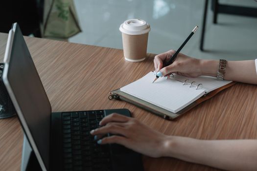 Closd up business woman taking note on paper with and she is using a laptop computer on a wooden desk in the office. Web banner.