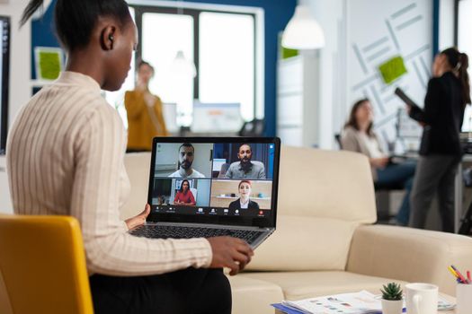 African manager woman discussing with remote colleagues using video call holding laptop sitting on couch in business modern office. Diverse coworkers planning new financial project in modern company