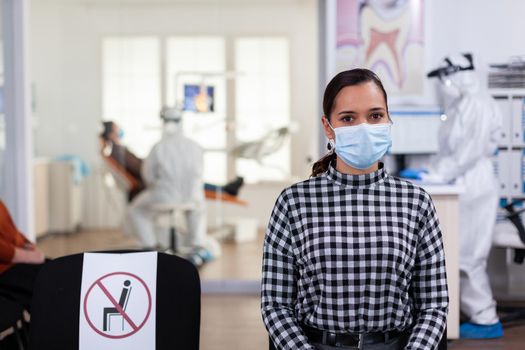 Portrait of woman in dental office looking on camera wearing face mask sitting on chair in waiting room clinic while doctor working. Concept of new normal dentist visit in coronavirus outbreak.