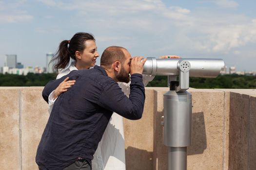 Happy couple enjoying summer spending time on building tower enjoying looking at landscape with metropolitan city using binoculars telescope. Panorama with urban buildings from observation point