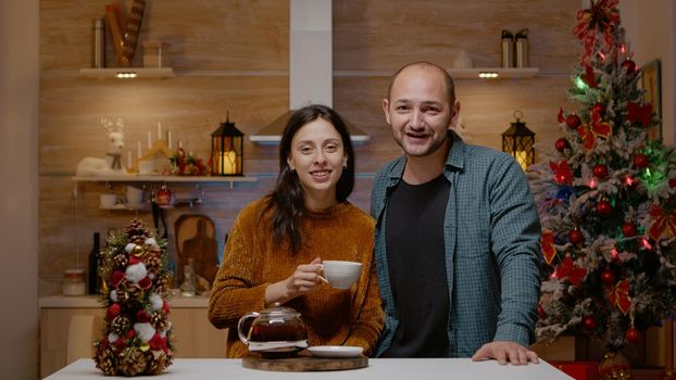 Couple using video call for festive communication with family at christmas time. Married man and woman talking on online conference for holiday season celebration in decorated kitchen.