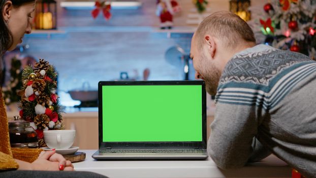 Couple using horizontal green screen on christmas eve looking at laptop. Man and woman watching chroma key with isolated background and mockup template celebrating holiday festivity.