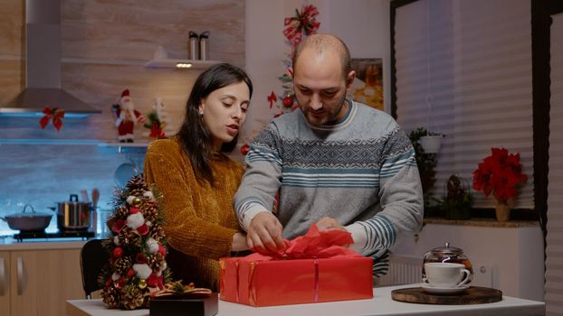 Couple preparing gift box for family on christmas eve festivity. Festive man and woman wrapping paper and ribbon on present for seasonal celebration and winter holiday. People decorating