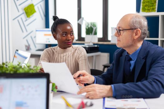 African employee discussing with senior executive looking financial charts in startup company conference board room. Multiethnic businesswoman and mature entrepreneur brainstorming.