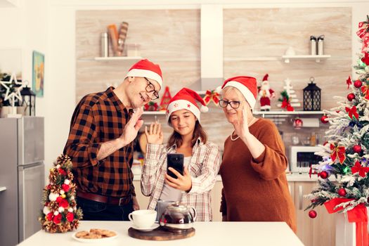 Niece and grandparents celebrating christmas saying hello at phone in time of video conference with relatives. Happy multi generation family wearing santa hat during video conference celebrating winter holidays, decorated kitchen.