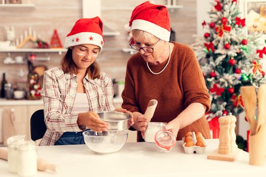 Multigeneration family doing cookies together on christmas day. Happy cheerful joyfull teenage girl helping senior woman preparing sweet buiscuits to celebrate winter holidays wearing santa hat.
