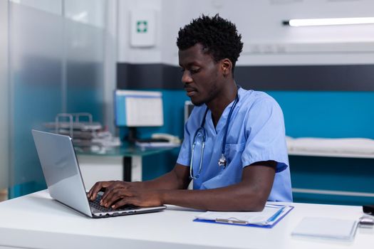 Black young man working as nurse at medical clinic wearing uniform and stethoscope. African american person sitting at desk while using modern laptop, typing on keyboard in cabinet