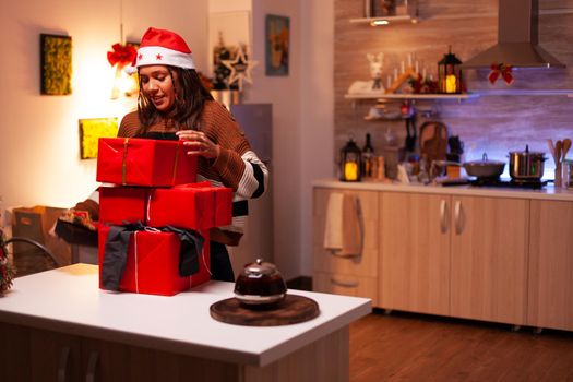 Modern adult moving present boxes on counter at festive home. Caucasian young woman wearing santa hat holding gifts for friends and family at christmas eve celebration holiday festivity