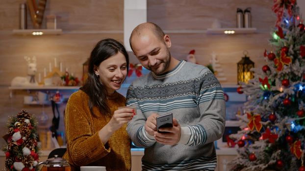 Festive couple buying presents with credit card on smartphone, doing online shopping for christmas eve celebration. People using device paying for gifts in seasonal decorated kitchen