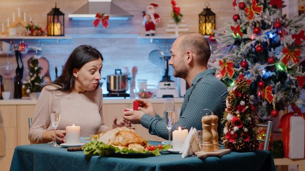 Loving couple enjoying festive dinner with traditional food in decorated kitchen. Man with engagement ring proposing to woman on christmas eve feeling happy. Romantic holiday celebration