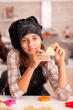 Portrait of grandchild wearing apron looking into camera while holding cookie dough using baking shape preparing gingerbread dessert. Child celebrating christmas season in xmas decorated kitchen